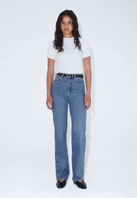 Bílý top s krátkými rukávy spárovaný s modrými džíny s vysokým pasem. Úzký střih, světlá modrá džínovina, rovný střih nohavic, doplněno černým páskem a černými botami.