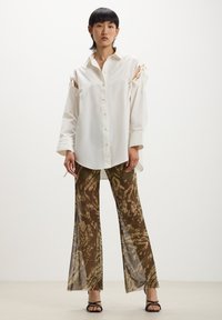 Camicia bianca con scollature sulle spalle e nastri, abbinata a pantaloni larghi a fantasia marrone con toni e texture terrose.