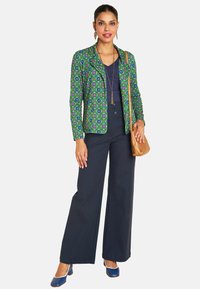 Patterned green blazer with floral design, navy top, high-waisted navy trousers, and blue heels. Gold handbag and accessories included.