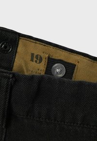 Juodi džinsai su matoma ruda juosmens juosta ir etiketė, kuriame yra skaičius "19". Yra sidabrinė sagtys ir tekstūruotas audinys.