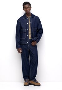 Giacca di jeans con tasche frontali, abbinata a una camicia beige a righe e pantaloni di jeans larghi. Scarpe marroni con texture e lacci blu.