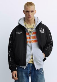 Zwarte bomberjack met witte accenten, gedragen over een grijze hoodie. Gestreept geel-grijs T-shirt eronder. Blauwe spijkerbroek met kettingdetail.