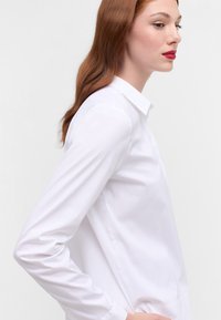Camicia bianca a maniche lunghe con bottoni, caratterizzata da un colletto a punta, tessuto liscio e vestibilità su misura, mostrata di lato su uno sfondo semplice.