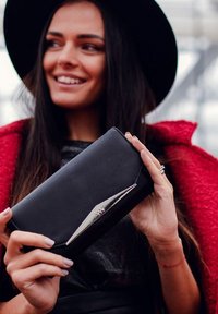 Black leather wallet with a sleek design, featuring a silver metal accent on the front. Held in hands against a red coat backdrop.