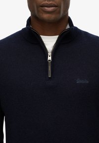 Granatowy sweter zapinany na zamek z ryflowaną strukturą, z kołnierzykiem zapinanym na stojąco oraz małym haftowanym logo "SuperDry" na klatce piersiowej.