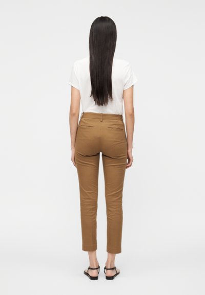 Pruunid keha järgivad püksid, millel on sile tekstuur, tagataskud ja pahkluuni ulatuvad sääred, kombineeritud lihtsa valge t-särgiga.