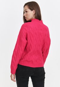 Różowy sweter w warkocze z wysokim kołnierzem i ściągaczem. Posiada długie rękawy i fakturowany wzór. Noszony z czarnymi spodniami.