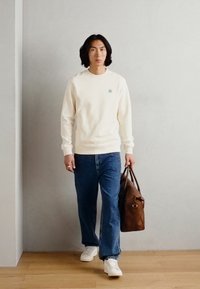 Crème sweatshirt met klein groen logo, blauwe jeans, witte sneakers en een bruine leren tas, staand op een houten vloer tegen een grijze muur.