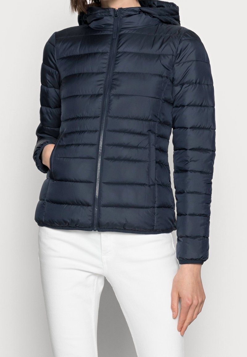 Personne portant une veste matelassée à capuche bleu foncé avec une fermeture éclair à l'avant et un pantalon blanc, une main dans la poche et l'autre détendue sur le côté.