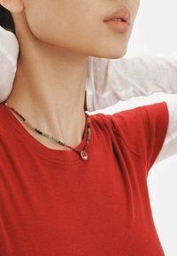 Collar de cuentas con varios colores, que presenta un colgante de gema central, llevado sobre una camiseta roja de cuello redondo.