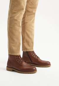Brązowe skórzane botki za kostkę o gładkim wykończeniu, okrągłym nosku, sznurowanym przodzie i bocznym zamku. Gumowa podeszwa, w zestawieniu z beżowymi spodniami z kordurowej tkaniny.