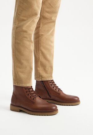 Brązowe skórzane botki za kostkę o gładkim wykończeniu, okrągłym nosku, sznurowanym przodzie i bocznym zamku. Gumowa podeszwa, w zestawieniu z beżowymi spodniami z kordurowej tkaniny.