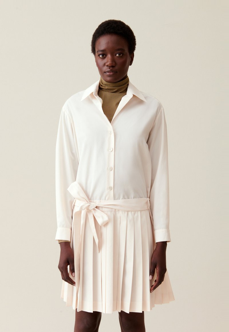 Robe chemise à manches longues en tissu ivoire doux avec une taille nouée, dotée d'une jupe plissée et d'un col structuré. Sous-pull à col roulé texturé en olive.
