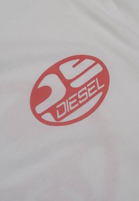 T-shirt en coton blanc arborant un bold logo circulaire rouge avec le mot "DIESEL" dans une police moderne et des éléments de design abstraits.
