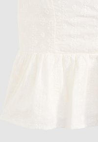 Top en coton blanc avec broderie florale texturée et ourlet en volants évasés, présentant une surface lisse et un design de corsage ajusté.