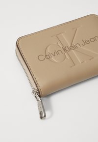 Bežinė dirbtinės odos piniginė su iškilniu "Calvin Klein Jeans" logotipu, užtrauktuko uždarymu ir sidabriniais detalėmis. Lygus tekstūra ir stačiakampio forma.