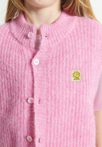 Roza pletena brezrokavnik z rebrasto teksturo, ki ima okrogel ovratnik, sprednje gumbe in majhen logotip cveta. Mehka, topla materiala.