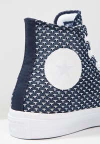 Laivastonsininen korkeavarpainen lenkkari, jossa on valkoinen tähtilogo, neulottu kuvioitu kangas, kumipohja ja valkoiset aukot. Ommellut saumat reunojen ympäri.