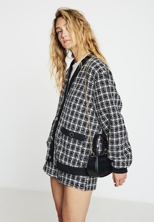Veste en tweed noir et blanc à coupe décontractée, dotée de deux poches avant et de poignets côtelés. Un petit sac à bandoulière noir est porté.