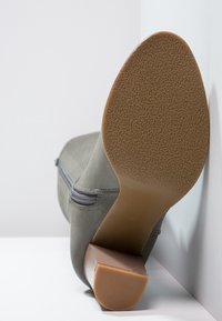 Botte en daim gris avec une tige lisse, une fermeture éclair sur le côté et une semelle en caoutchouc texturée. Le talon est beige, offrant un contraste de couleur.