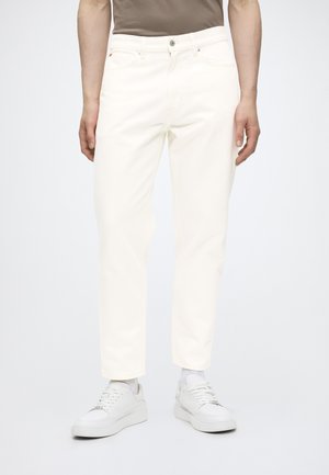 Personne portant un jean droit écru, des chaussettes blanches et des baskets blanches, debout devant un fond uni.