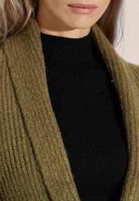 Olivgrüner, gestrickter Cardigan mit strukturiertem Design, über einem taillierten schwarzen Ripp-Turtleneck getragen. Nahaufnahme des Stoffes und der Farben.