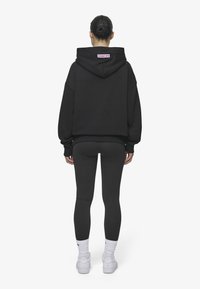 Hanorac negru oversized cu glugă și manșete cu nervuri, purtat cu colanți negri și adidași albi. Hanoracul are un logo pe spate.