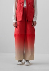 Rote, weit geschnittene Hosen mit einem Ombre-Effekt, der zu Beige verblasst, aus einem glänzenden Material. Kombiniert mit einem passenden ärmellosen Weste.