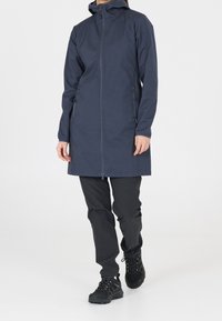 Marinblå lång jacka med dragkedja, justerbar huva och dragkedjeförsedda sidofickor. Kombinerad med svarta byxor och robusta svarta skor.