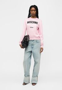 Femme aux longues tresses portant un pull rose Moschino Milano, un jean bleu clair ample, des sandales noires et tenant un sac à main noir.