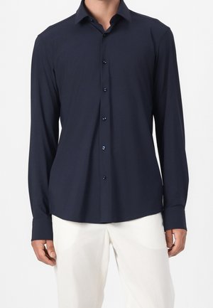 Camicia blu navy a maniche lunghe con bottoni, indossata con pantaloni bianchi su una persona in piedi su sfondo semplice.