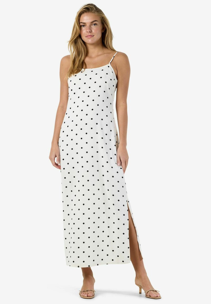 Femme portant une robe maxi blanche sans manches avec des pois noirs et une fente sur le côté, associée à des sandales beiges à talons.