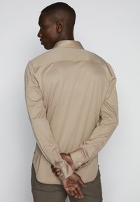 Beige Langarmhemd mit Kragen, glatter Textur und geknöpften Manschetten. Die Rückseite hat eine Schulterpasse und einen leicht abgerundeten Saum.