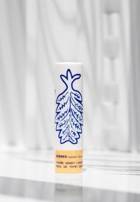 Vit cylindrisk läppbalsamsbehållare med blått lövmönster. Guldaccent vid basen med produktnamn och smaktext i svart.