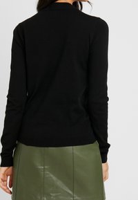 Pull en maille noire à manches longues et encolure ronde, avec une texture douce et des poignets côtelés, associé à une jupe en cuir verte.