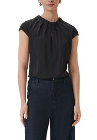 Sort kortærmet bluse med plisseret hals, der har en subtil nøglehulåbning. Glat stof med en afslappet pasform, sammen med mørkeblå jeans.