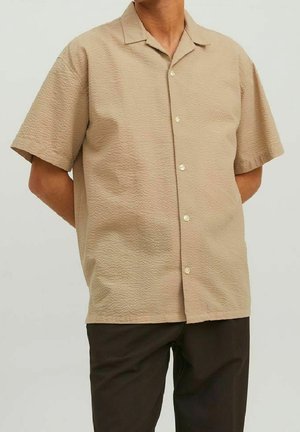 Homme portant une chemise boutonnée à manches courtes beige texturée et un pantalon marron foncé, les mains derrière le dos.