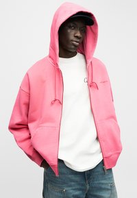 Ung mand iført en lyserød hættetrøje med lynlås og hætte oppe, hvid T-shirt, blå jeans og en sort kasket, stående med hænderne i lommerne.