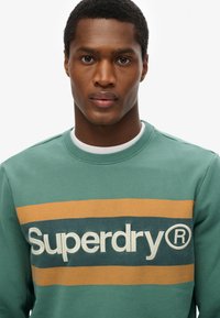 Zielona bluza z okrągłym dekoltem, z dużym logo "Superdry" w kremowym kolorze na pomarańczowo-granatowym tle w paski. Miękka tkanina.