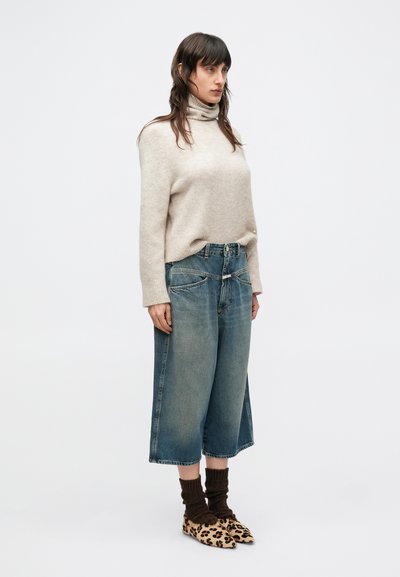 Pull beige à col roulé tricoté, culottes larges en denim clair, chaussettes marron côtelées et chaussures à imprimé léopard, vue de profil avec un fond neutre.