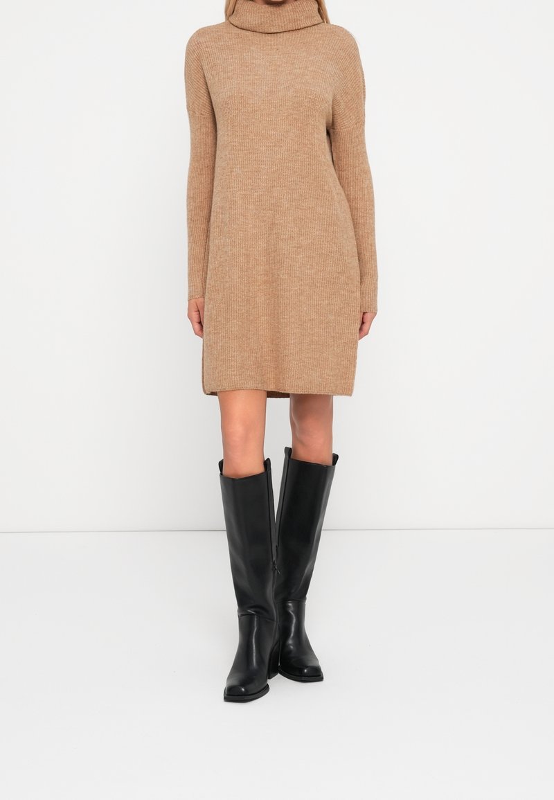 Vestito in maglia a coste beige con collo alto, maniche lunghe e vestibilità comoda, abbinato a stivali alti in pelle nera.