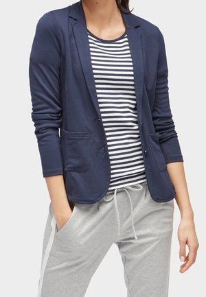 Femme portant un blazer bleu marine sur une chemise rayée noir et blanc et un pantalon gris clair à cordon, les mains dans les poches.