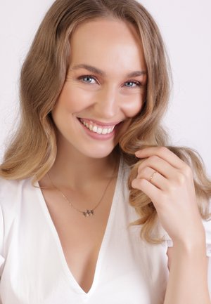 Goldene Halskette mit drei kleinen Anhängern. Das Model trägt ein weißes Oberteil, hat lange, wellige Haare und einen zarten goldenen Ring an ihrem Finger.