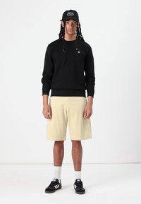 Sort sweatshirt med ribbede manchetter, lysebeige shorts og sorte sneakers. Kasketten har tekst. Modellen står foran en ensfarvet baggrund.