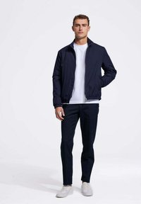 Navy bomberjack met een rits, elastische mouwen, gecombineerd met een wit T-shirt en donkere broek, gedragen met lichte sneakers op een effen achtergrond.