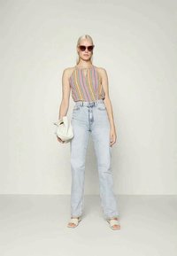 Top sem mangas com padrão colorido, jeans de cintura alta azul claro, sandálias brancas e uma bolsa branca. Modelo com cabelo loiro liso e óculos de sol.