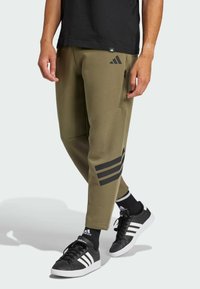 Olijfgroene joggingsbroek met ritszakken, met een zwart drie-stripes logo op het linkerbeen. Gecombineerd met zwarte sneakers en sokken.