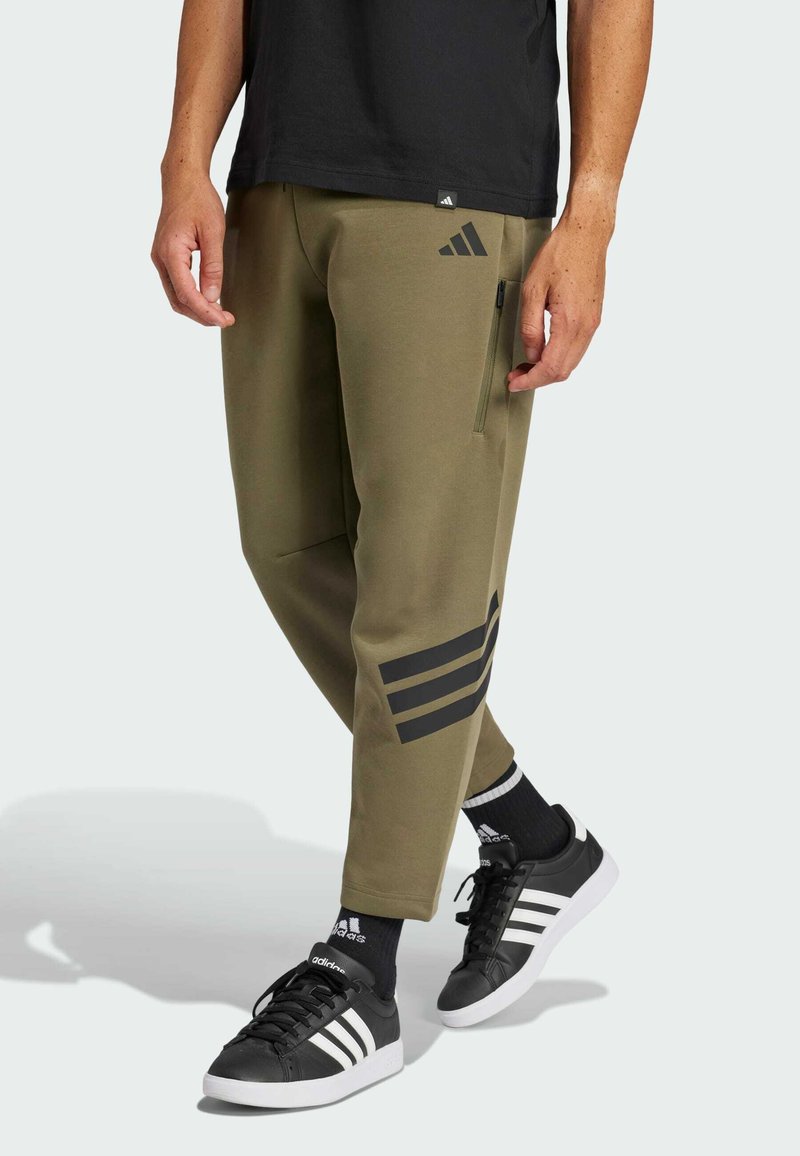 Olijfgroene joggingsbroek met ritszakken, met een zwart drie-stripes logo op het linkerbeen. Gecombineerd met zwarte sneakers en sokken.