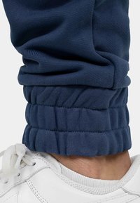 Marineblauwe sweatpants met opgerolde boorden, gemaakt van gestructureerd katoen, gecombineerd met een witte sportschoen met veters en een glad oppervlak.