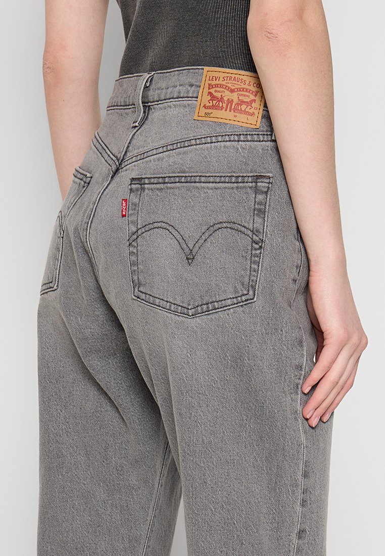 Vaqueros Levi's 501 grises con bolsillos traseros, una etiqueta roja de Levi's y un parche de piel marrón de la marca, llevados por una persona con una camiseta negra.